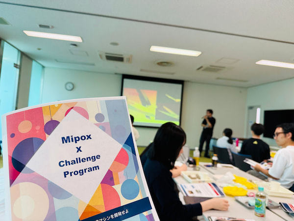 会場に配布された「Mipox X Challenge Program」の冊子。奥では社員と高校生がオリエンテーションに耳を傾けます。